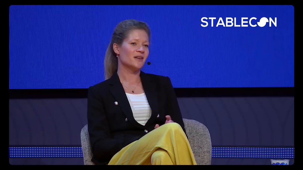The image shows a woman sitting in a chair with her hands on her lap while talking on a microphone. The background features a blue screen displaying white text that reads "@Visa's latest announcement on AI commerce, tokenized infrastructure, and stablecoin settlement reflects one of the biggest themes shaping financial infrastructure today: convergence". This suggests that the image is related to an event or discussion at Stablecon EMEA where the woman might be speaking about the current trends in financial technology.