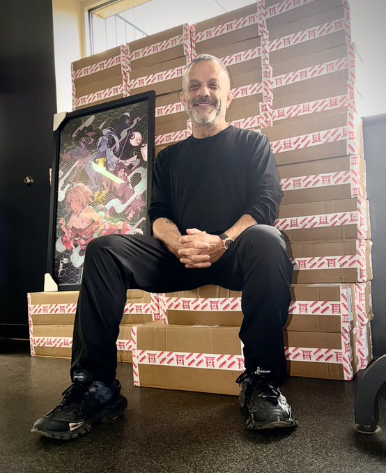 A man with a gray beard, wearing a black long-sleeve shirt, black pants, and Balenciaga sneakers, sits smiling on a stack of cardboard boxes sealed with red and white "Azuki" branded tape, with a framed anime-style artwork beside him. The image directly illustrates the post's announcement of receiving 7,200 Azuki TCG booster packs, with the man surrounded by the massive haul in preparation for an upcoming community party.