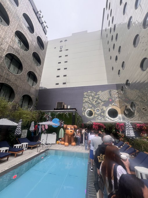 An outdoor pool area surrounded by unique buildings with circular windows, where a large brown bear statue sits near the edge as people gather around.