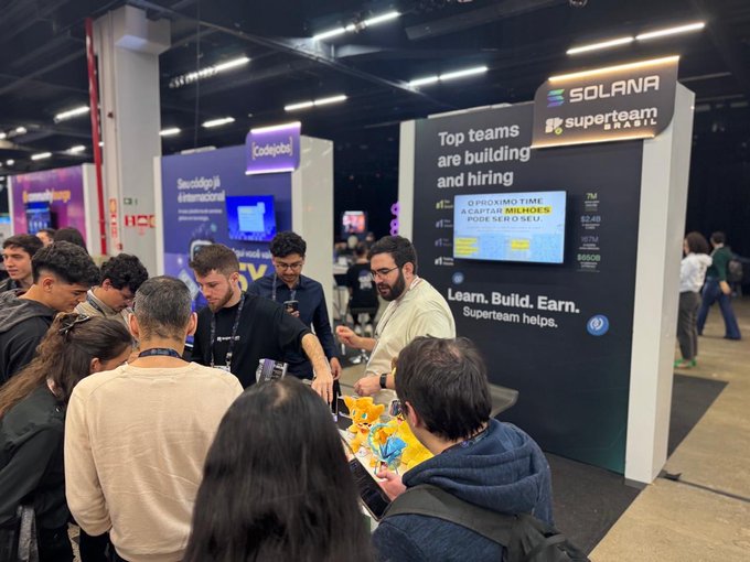 A crowd gathers at a Solana Superteam Brazil booth at what appears to be a tech conference, with a large screen displaying 'Top teams are building and hiring' and 'Learn. Build. Earn. Superteam helps.'