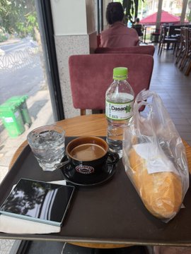 A cafe table scene featuring a bottle of Dasani water, a cup of coffee, a glass of water, a bread roll in a plastic bag, and a menu or book, with a view of outdoor seating.