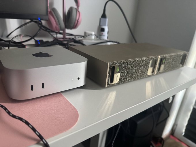 An Apple Mac mini sits beside a textured, box-shaped device on a white desk shelf, with the Mac's logo and the device's "DGX" label visible. This setup illustrates the post's humor of "doubling" the hardware by adding two more DGX Sparks to the workstation alongside the existing Mac mini.