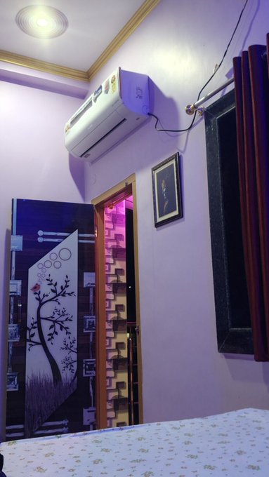 An interior room with lavender walls, featuring a wall-mounted air conditioner, a decorated wooden door with a tree design, a framed portrait, and dark red curtains beside a window.