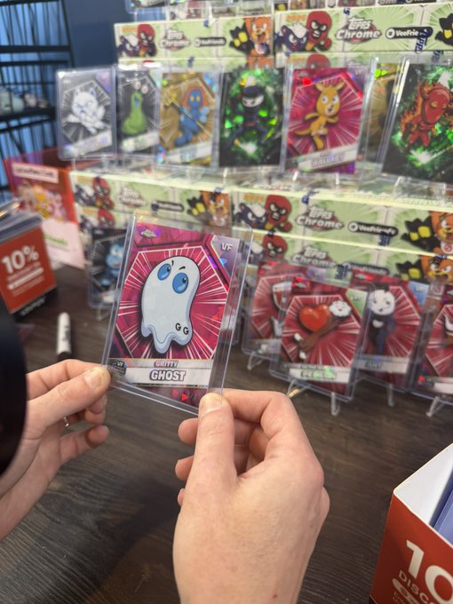 Hands hold up a Topps Chrome 'Gooey GHOST' trading card in a store, with a display of other character cards visible in the background.