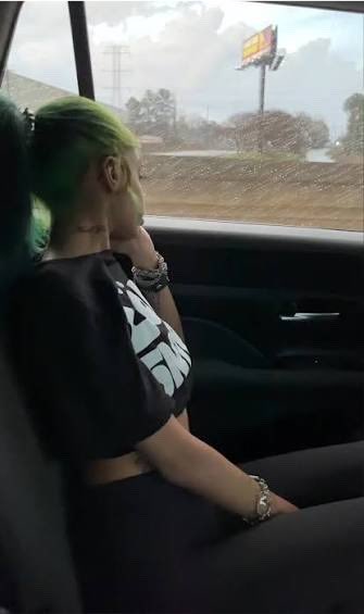 A person with bright green hair, wearing a black 'STAR WARS' t-shirt and bracelets, sits in the passenger seat of a car with their hand near their face, looking out the window at a rainy landscape with a billboard.