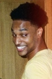 Casual photo of a smiling young Black man with a short fade haircut, wearing a tan shirt against a plain wall.