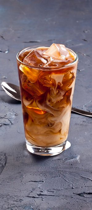 A tall glass of iced coffee with swirling cream sits on a dark, textured surface next to a silver spoon.