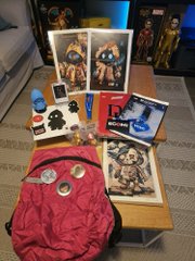 A collection of anime merchandise and figurines is displayed on a wooden table, including framed artwork, a red backpack, and various collectible figures.