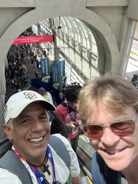 Two smiling men wearing sunglasses and event lanyards pose for a selfie on an escalator, with a crowded convention hall visible in the background.