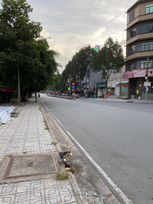A quiet city street scene features a tiled sidewalk, a green-canopied 'GÀ VÀNG' storefront, traffic lights showing green, and a few distant motorbikes under a cloudy sky.