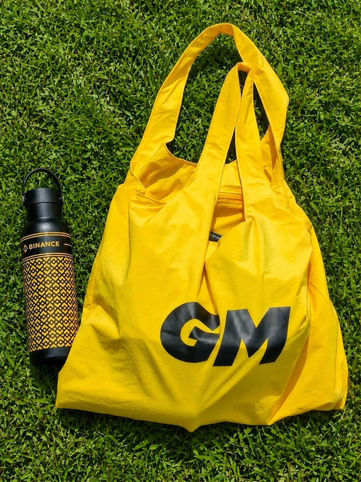A bright yellow 'GM' (Good Morning) reusable bag lying on grass next to a black and gold Binance water bottle.