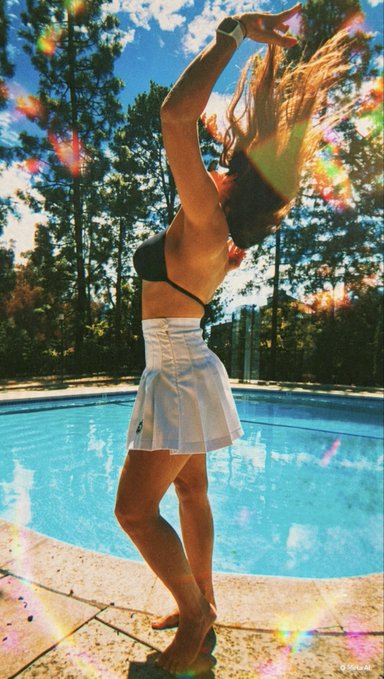 A young woman in a black top and white pleated skirt stands barefoot by a swimming pool, stretching her arms overhead as sunlight creates rainbow lens flares.