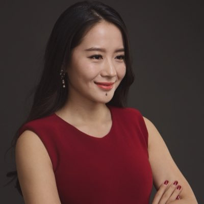 A woman with long dark hair wearing a red sleeveless top smiles confidently with her arms crossed, exuding poise against a dark background.

The image appears to represent one of the female crypto role models mentioned in the post, embodying the "stronger women in crypto" theme by showcasing a poised, professional woman who could symbolize the leadership and influence being celebrated.