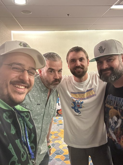 A group selfie of four smiling men at what appears to be a convention, wearing various hats and graphic t-shirts, with one shirt reading "TECHNOLOGY HUT".