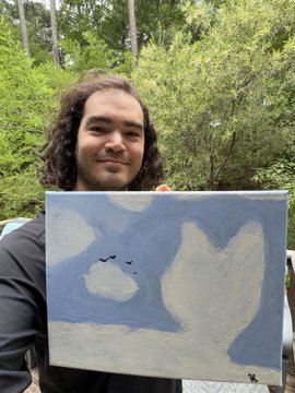A young man with curly hair standing outdoors in front of trees, proudly holding up a small canvas painting of a blue sky with white clouds and birds.