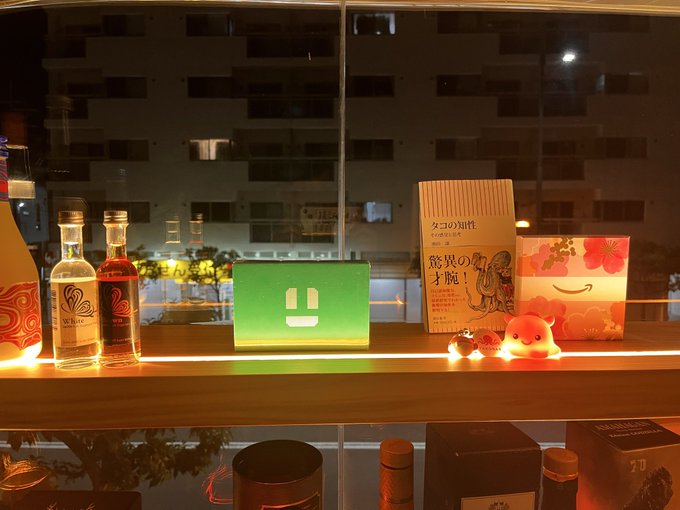 urns, bottles, and a book with Japanese characters are arranged on a shelf, illuminated by warm yellow light from the window behind them.