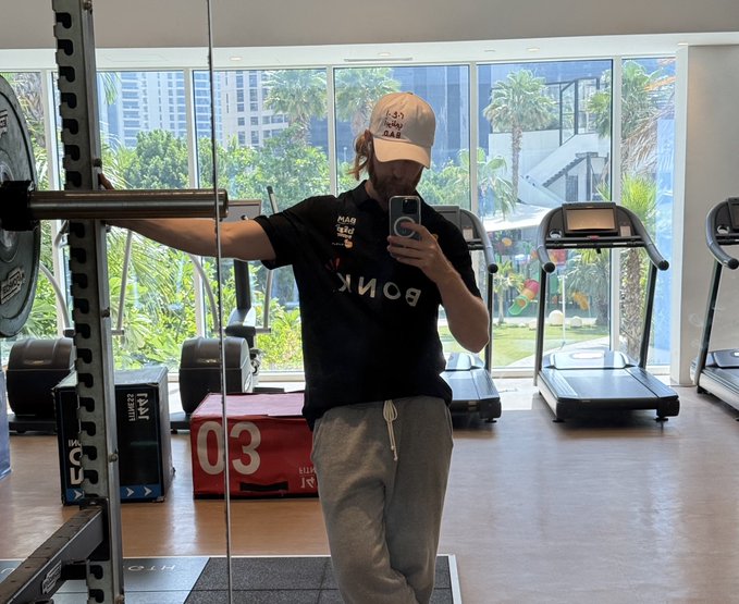 The image features a man standing in front of a gym, taking a selfie with his phone while wearing a baseball cap. The gym has various exercise equipment such as treadmills and weight machines scattered around the room. There are also several benches placed throughout the space for people to use during their workouts. A bench can be seen near the center of the image, while another is located towards the right side. Additionally, there's a bench on the left side of the gym area. The man appears to be capturing his time at the gym and possibly sharing it with others through social media platforms like Twitter or Instagram.