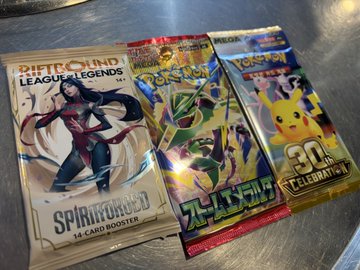 Three Pokémon trading card game booster packs, including 'Spiritmasked' for League of Legends, Japanese 'Pokémon Card Game' featuring Rayquaza, and a 'Celebrate' 30th anniversary pack featuring Pikachu, resting on a metallic surface.