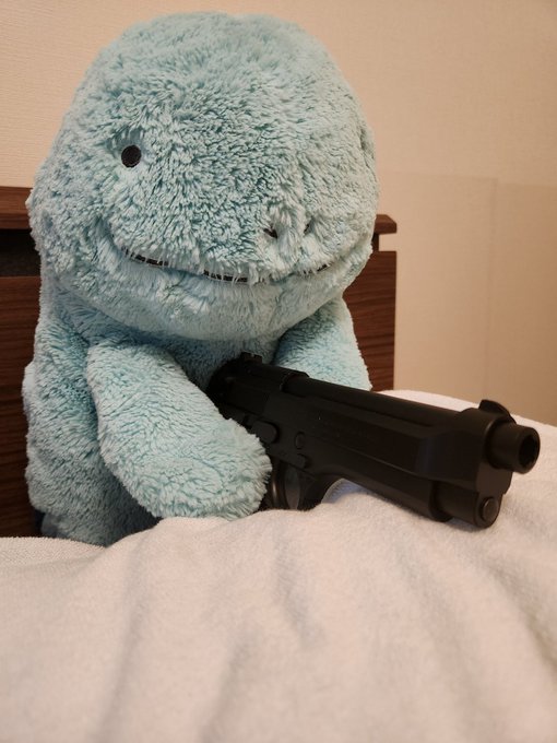 A light blue plush dinosaur toy sits in a bed, holding a black handgun in its paws.