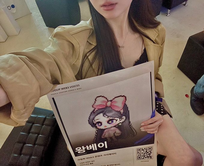 A young woman wearing a tan leather jacket and a necklace holds up a fan sign featuring a cartoon girl with a pink bow, with Korean text and 'JOUJ WEB VOICES' visible along with a QR code.