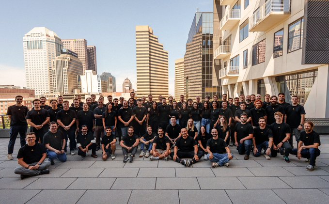 A large group of employees wearing matching black t-shirts poses for a team photo on an outdoor rooftop terrace, with a city skyline featuring tall buildings visible in the background.