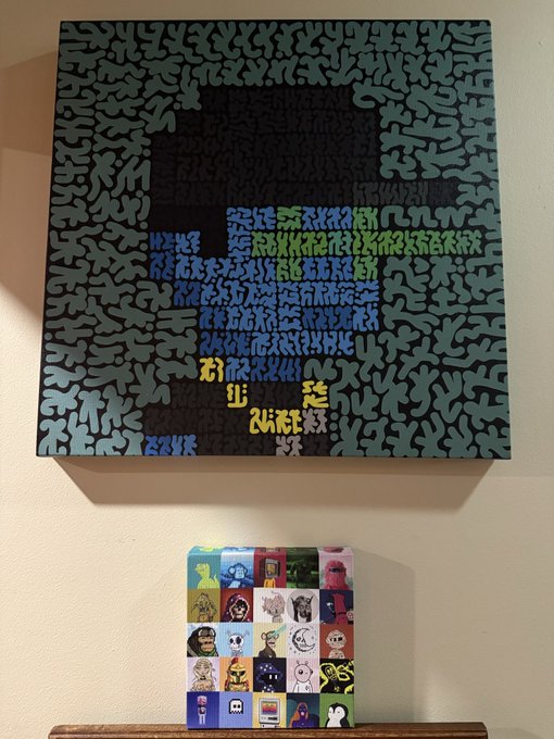 A square canvas featuring abstract, colorful glyph-like patterns hangs on a wall above a small box displaying a grid of 20 distinct cartoon-style character portraits, with one spot empty likely awaiting the user's new PFP. The artwork displays text fragments including "KOWSKY" along the top edge, reflecting the artist credit mentioned in the caption.