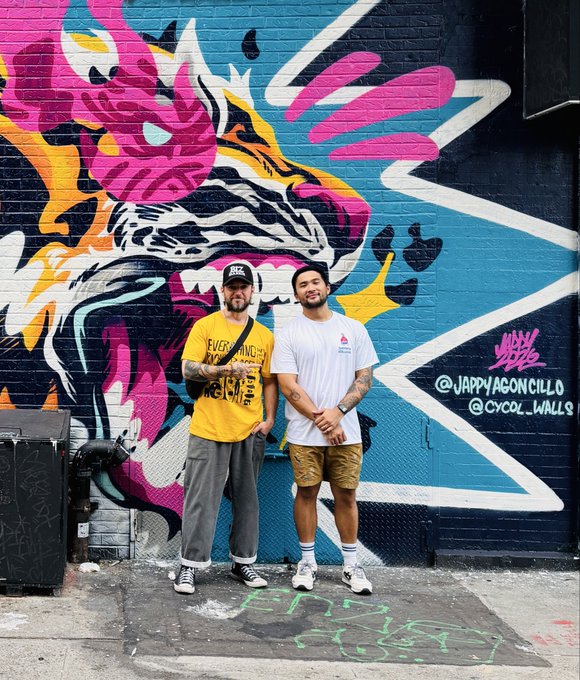 Two men pose in front of a colorful street art mural featuring a tiger-like face and pink hand; graffiti reads "@JAPPYAGONZILLO" and "@CYCOL_WALLS".