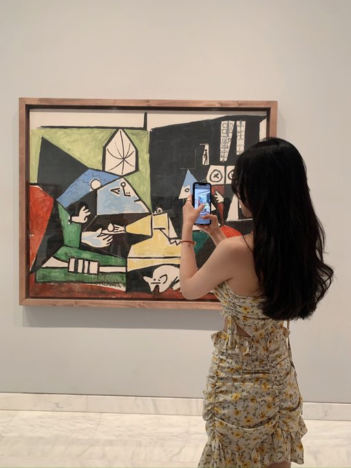 A woman in a floral dress takes a photo with her phone of Picasso's Guernica painting displayed in a gallery.