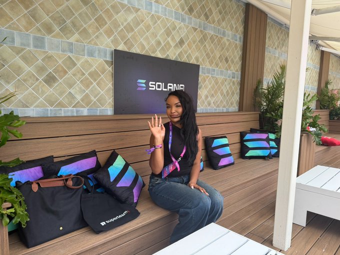 A woman wearing a lanyard sits on a wooden bench at a Solana-branded event space, waving with her hand next to Solana-themed merchandise bags and pillows.