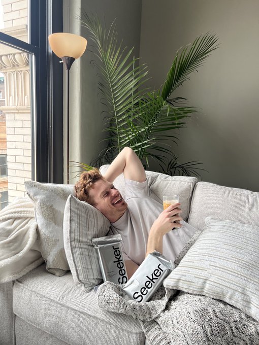 The image shows a man lying down on a couch with his arms raised, holding two boxes of Seeker rings. The man appears to be smiling while laying on the couch, possibly enjoying some leisure time or taking a break from work.