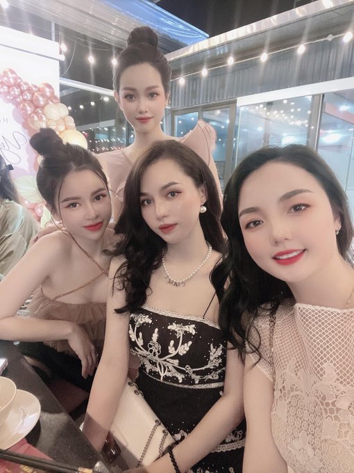 Four elegantly dressed women pose together for a selfie at what appears to be a celebratory indoor event with string lights and balloon decorations, with the woman in the center wearing a black embellished dress and pearl necklace. The festive gathering matches the caption's cheerful weekend greeting and wishes for a "radiant" day with good outcomes ahead.