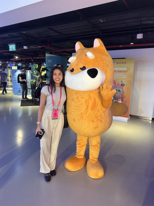 A young woman with a pink lanyard poses for a photo next to a large orange Shiba Inu mascot costume at what appears to be an indoor event or convention center.