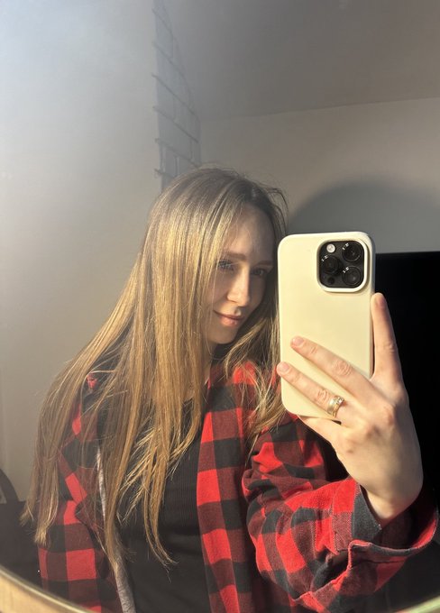 A mirror selfie shows a young woman with long blonde hair wearing a red and black plaid flannel shirt over a black top, holding a phone with a beige case.