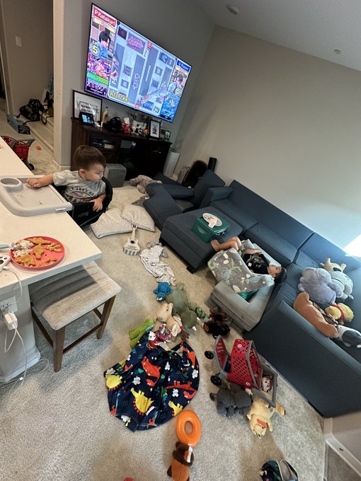 A living room scene shows two young children playing amid scattered toys, pillows, and blankets on the floor in front of a couch and a large wall-mounted TV.