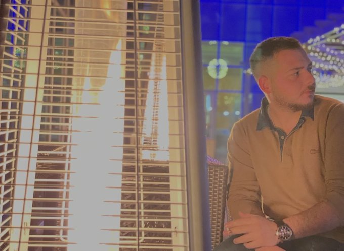 A man in a tan sweater sitting next to a tall outdoor patio heater, with blurred blue lights in the background.