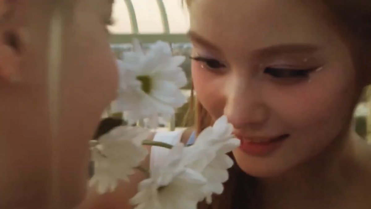 A close-up shot shows a young woman with shimmery eye makeup and a small tear delicately touching her face to white flowers in a sunlit setting.