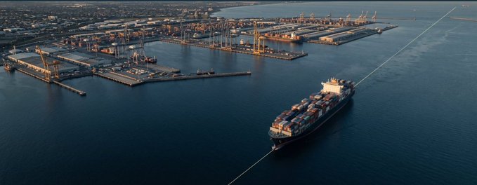 Aerial view of a large container ship loaded with cargo containers sailing past an industrial shipping port with cranes and docks.