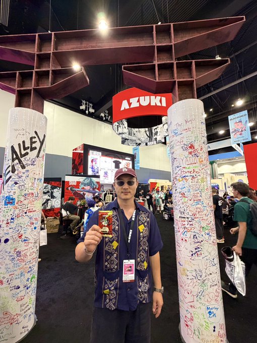 A man wearing a cap, sunglasses, and an Azuki lanyard stands between two signature-covered pillars at an Azuki convention booth, holding up a collectible while a large red "AZUKI" sign hangs above the busy expo floor. The image reflects the post's excitement about joining the Azuki community as a new supporter ("IKZ") witnessing the project's story unfold firsthand.