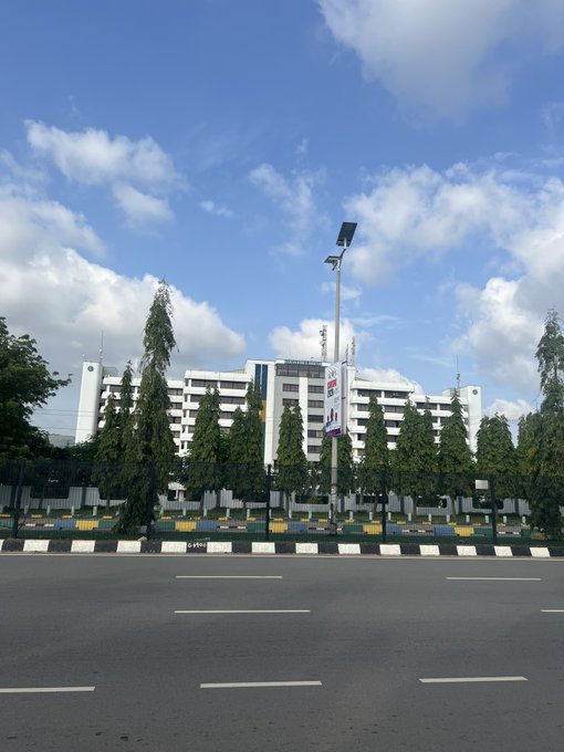 A wide, empty multi-lane road stretches across the foreground, bordered by a median with greenery and trees, leading to a large white commercial or office building complex under a bright blue sky with scattered clouds.