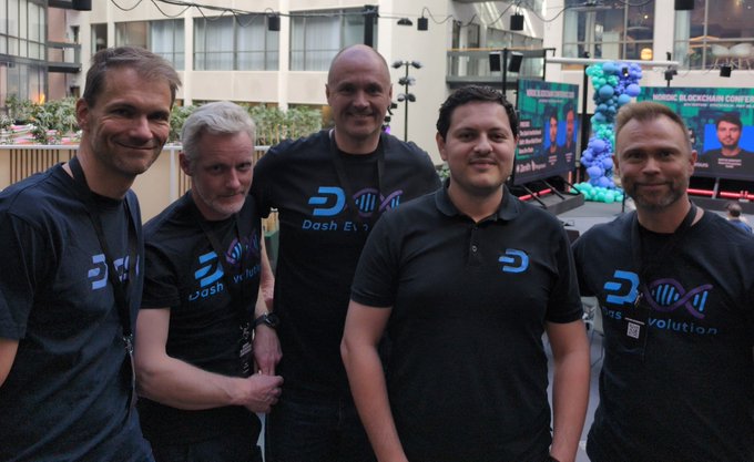 The image shows a group of three men wearing black t-shirts with blue text that says "Dash Growth", "@crowdnode", and "@DashInvests". They are standing together, posing for a photo at an outdoor event or conference. The background includes a building and balloons, suggesting the location is likely a public gathering or celebration.