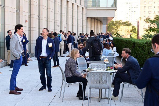 Two men in blazers chat on the patio of a modern building while others sit at a table with food, with a larger crowd mingling in the background at what appears to be a networking event.

The post's mention of cohosting the 6th Annual Fordham Law Blockchain Regulatory Symposium suggests this is a pre-event or post-event networking gathering for the symposium, where attendees and hosts like Donna Redel, Joyce Lai, and featured speaker Chris Giancarlo are connecting before or after the main agenda.