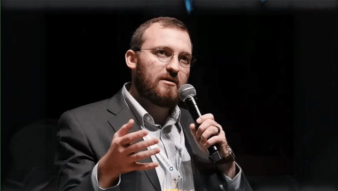 A bespectacled man with a beard, wearing a dark blazer over a checkered shirt, speaks into a microphone while gesturing with his hand, presumably Charles Hoskinson discussing Cardano's performance upgrades at a conference.