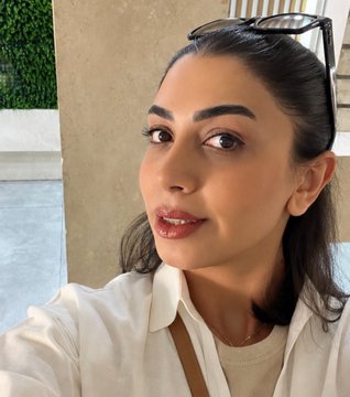 A close-up selfie of a woman with dark hair pulled back, sunglasses resting on her head, wearing a white shirt against an outdoor background.