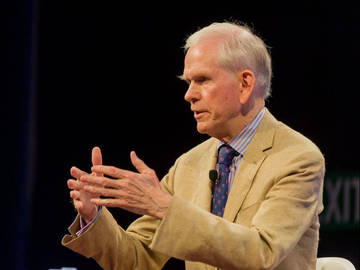 An older man with gray hair, wearing a tan jacket and blue patterned tie, speaks while gesturing expressively with both hands. The image accompanies a post about billionaire investor Jeremy Grantham's criticism of cryptocurrencies, with "KIN" partially visible in the background text.