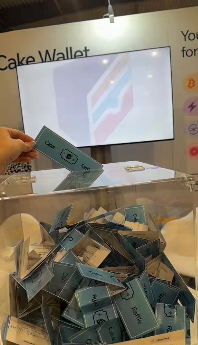The image shows a person holding a blue card with the word "cake" written on it, which appears to be a ticket or entry card for an event or competition. The person is standing next to a large pile of similar cards, indicating that there might be multiple events or competitions taking place at this location. The post also mentions a Bitcoin giveaway and encourages people to enter by stopping by the Cake Wallet booth at #BTCPrague.