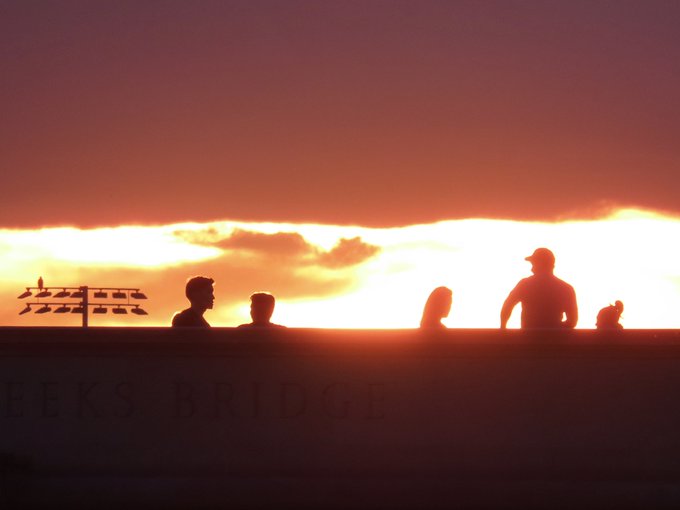 Silhouetted figures of people standing against a vibrant orange sunset sky, with 'EEKS BRIDGE' partially visible on a wall below and a wooden structure to the left.
