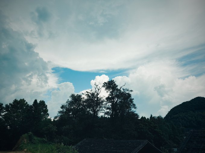 A scenic photograph showing a dramatic sky with large white clouds over a silhouette of trees and hills, with the tiled roofs of traditional houses visible in the foreground.