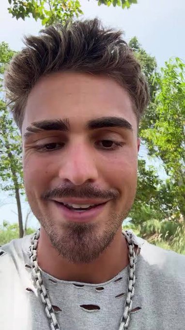 A close-up selfie of a smiling young man with dark hair and a light beard, wearing a grey distressed shirt and a silver chain necklace, outdoors with green trees in the background.