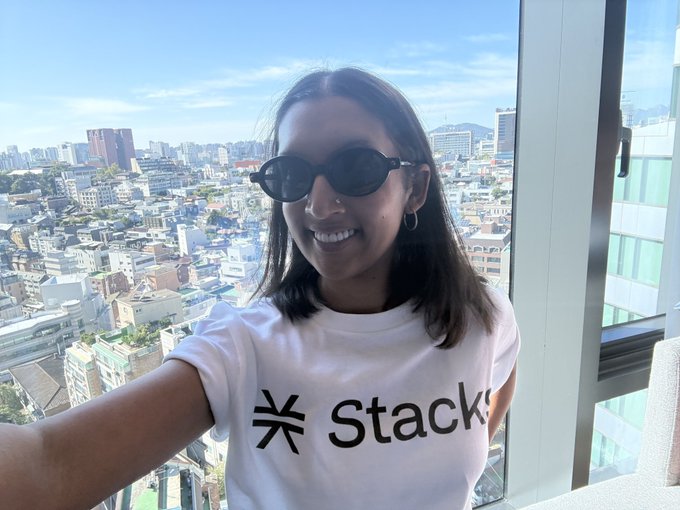 A young woman wearing sunglasses and a white "Stacks" t-shirt smiles for a selfie with an expansive cityscape visible through a window behind her.