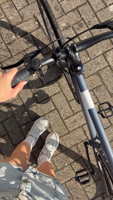 A first-person perspective photo showing a person's hand on the handlebar of a black bicycle, with their legs wearing denim shorts and white sneakers visible below on a brick paved surface.